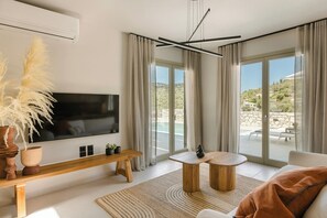 A modern living room with a flat-screen TV, wooden furniture, and large windows offering a view of a landscaped area. - Assos Reverie Villa (9GGR+RXG Erisos, Greece)