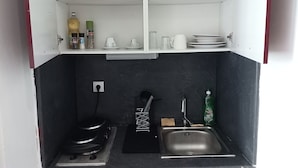 A compact kitchen with a stove, sink, and a shelf above holding various kitchen items. - Cosy Vallée Studio (Pau)