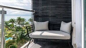 Un balcón con zona de estar acolchada, vistas a una piscina y palmeras, y una fachada de edificio moderna.