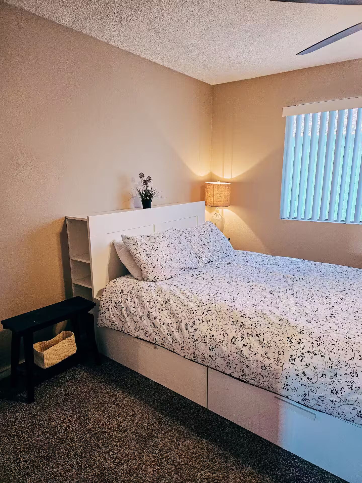 A bedroom with a bed, bedside table, lamp, and a window with blinds.