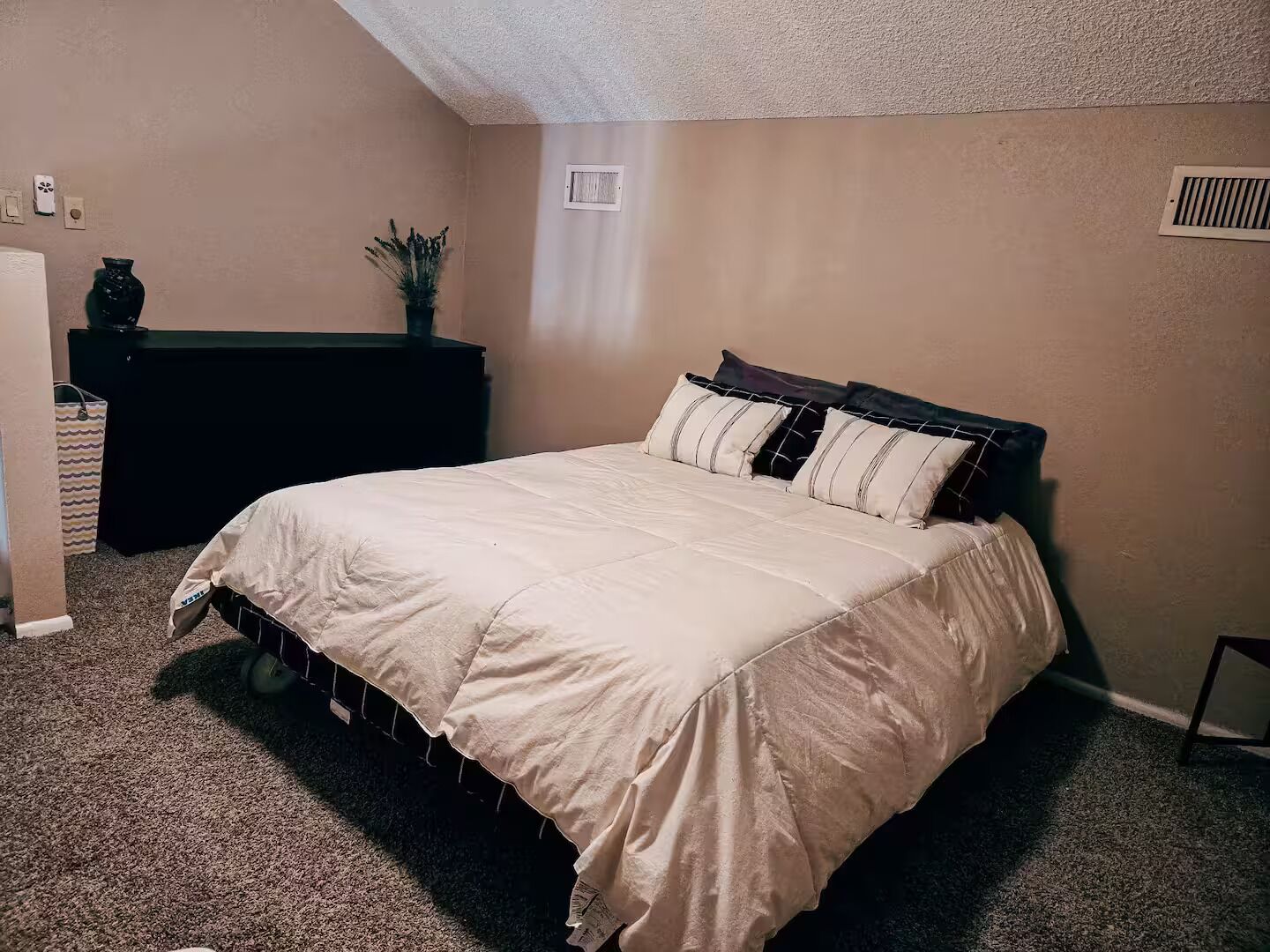 Une chambre à coucher avec un lit, une commode, une plante et une grille d’aération au mur.