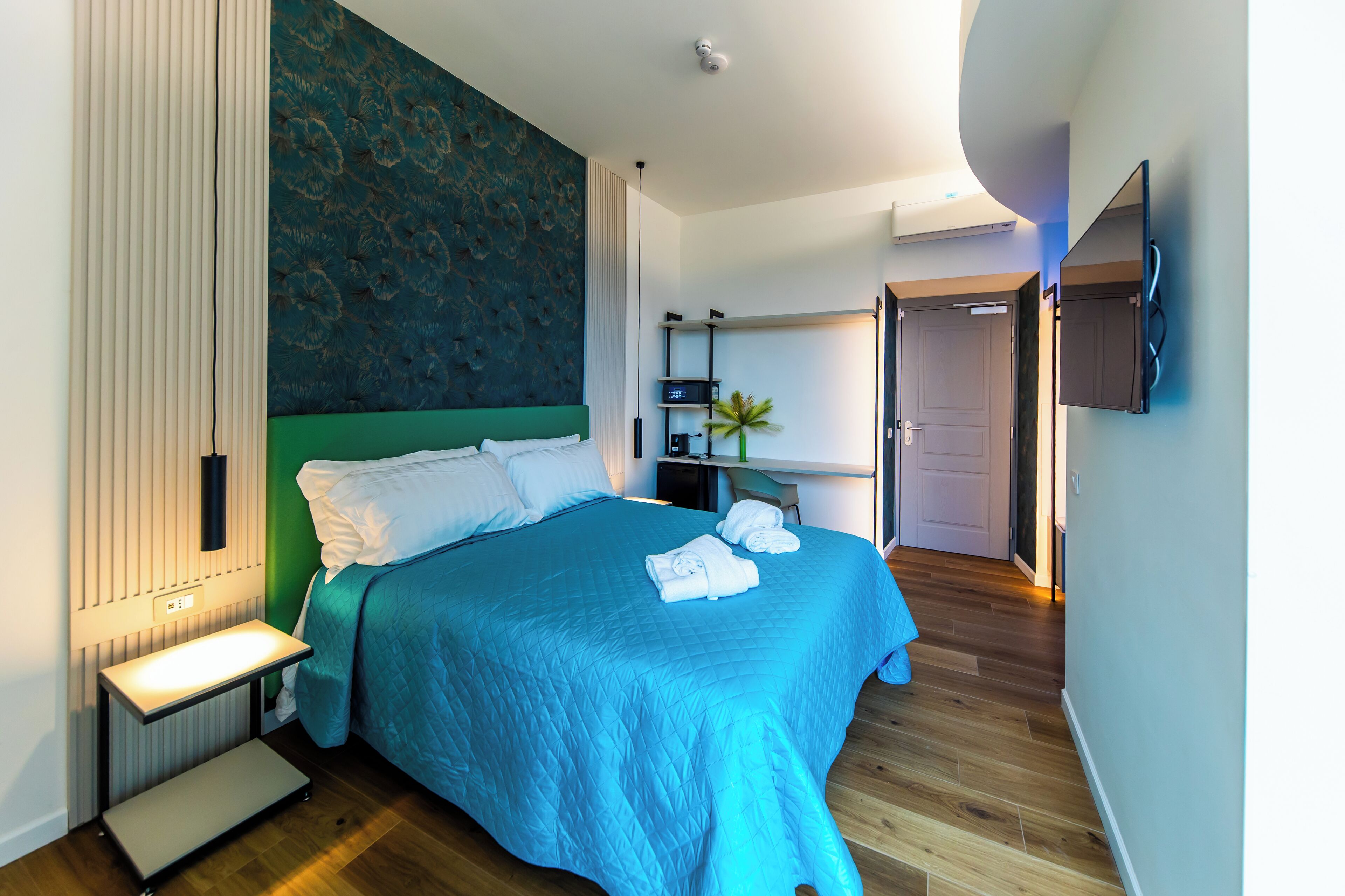A modern hotel room with a large bed, a bedside table, a flat-screen TV mounted on the wall, and a wooden floor.