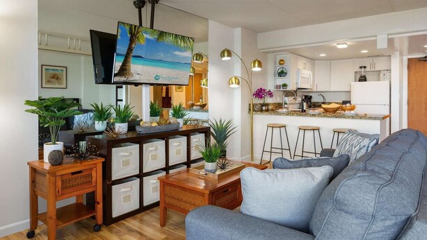 A modern living room with a large sofa, a wooden coffee table, and a TV mounted on the wall. There is a kitchen area with white cabinets and a bar counter. - 4th Fl Ilikai | Pool | AC | Marina View (Honolulu)