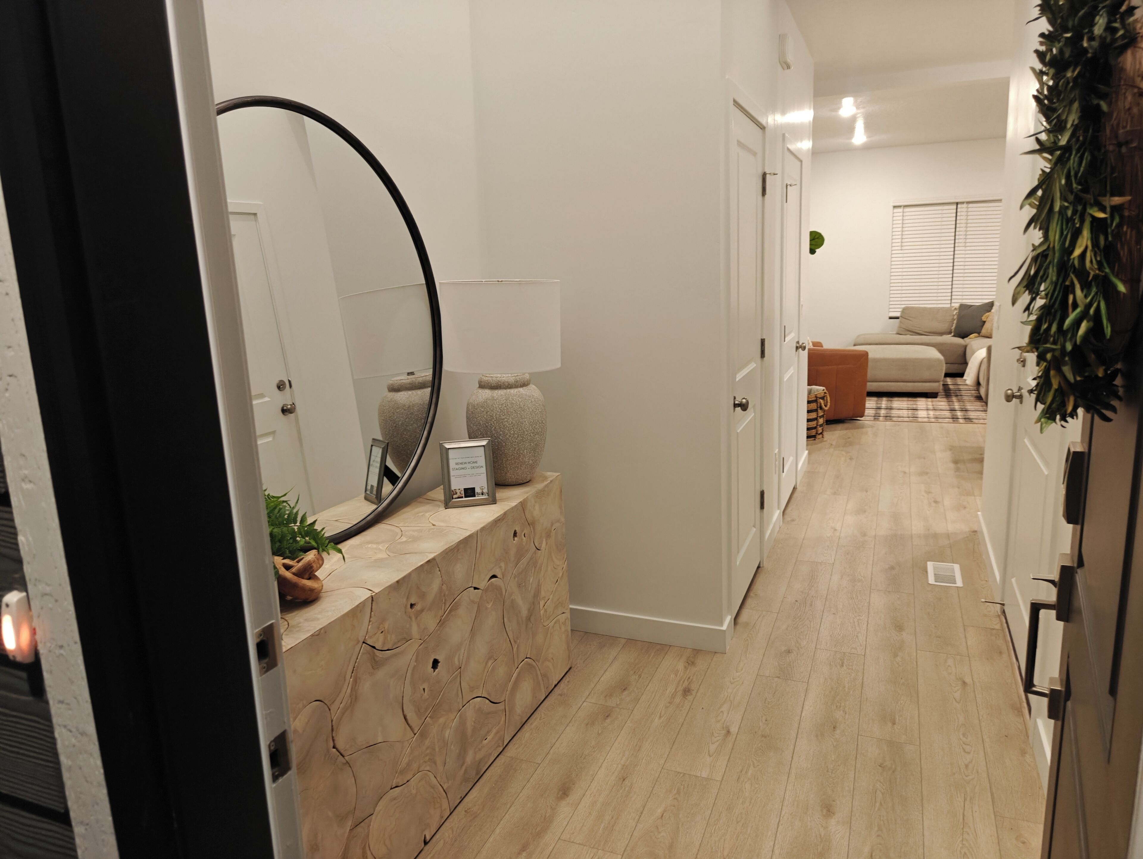Een hal met een houten console tafel, een grote spiegel en een lijst met een afbeelding. Een deur leidt naar een woonruimte met een bank en een plant.
