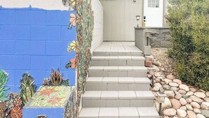 A set of concrete steps leading to a door, with a colorful mural on the left wall and a small garden area with rocks and plants.