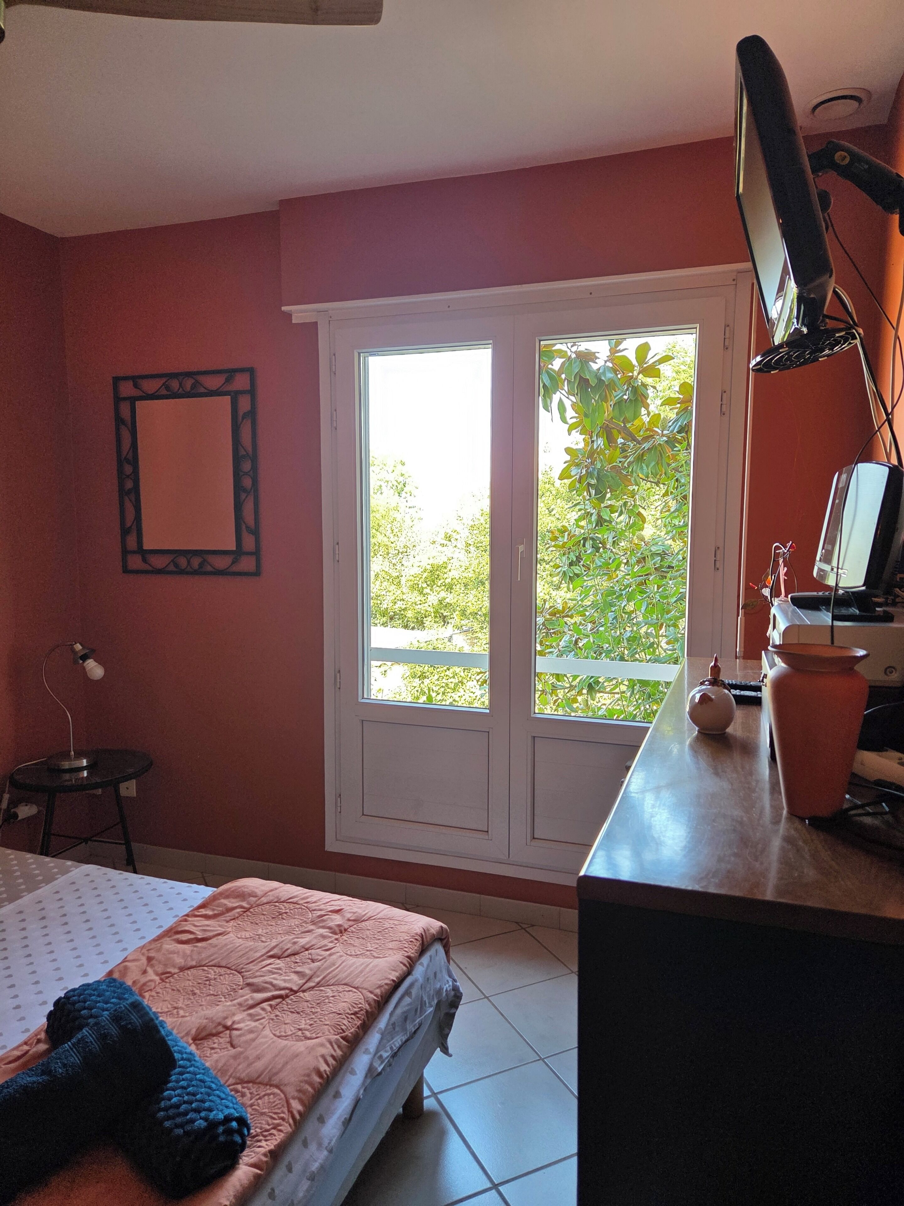 Een slaapkamer met een bed, een bureau met een computer, een spiegel en een raam met uitzicht op groen.