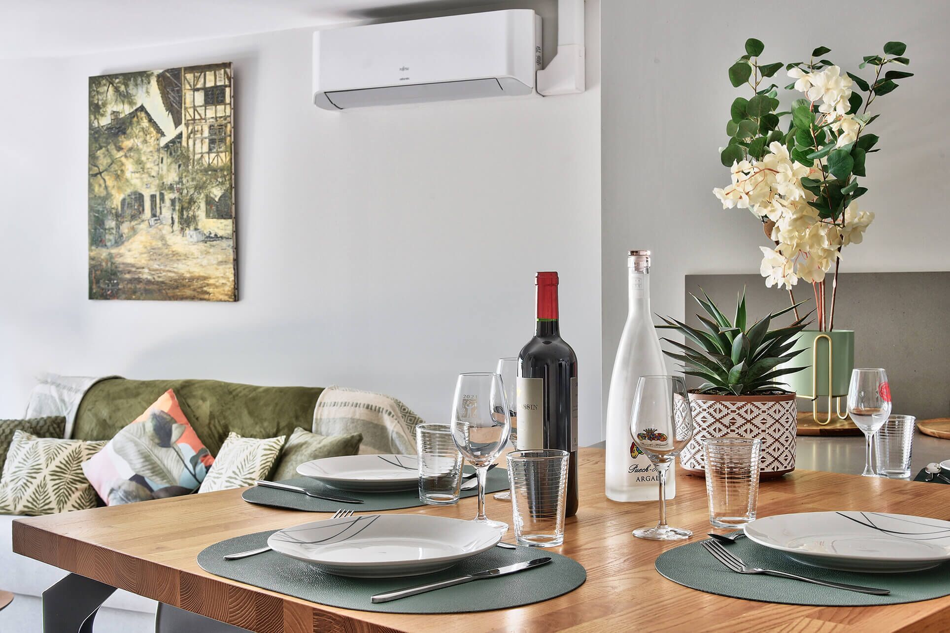 Un comedor con una mesa de madera dispuesta para dos personas, una botella de vino y una maceta con flores blancas.