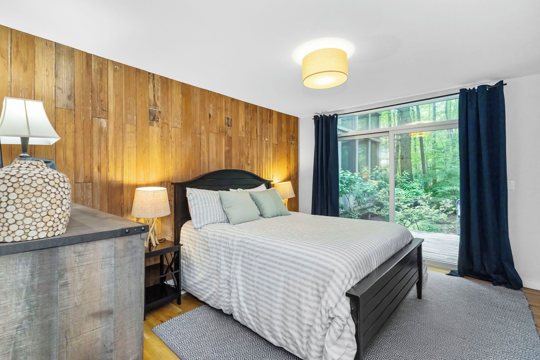 Une chambre avec un mur en bois, un grand lit, une table de chevet avec une lampe, et une vue sur la verdure par la fenêtre.