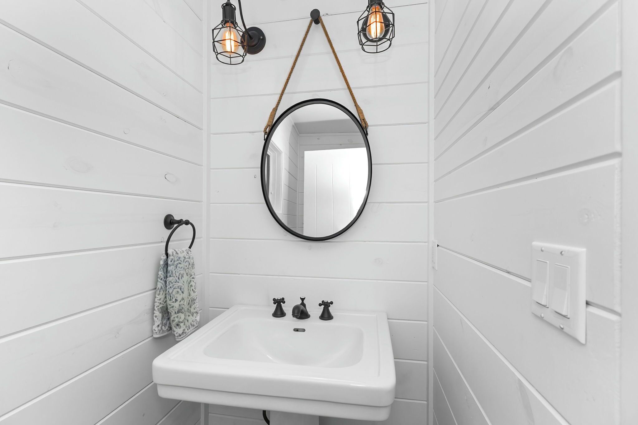 A bathroom with white wooden panel walls, a white sink with two faucets, a round mirror, and two wall-mounted light fixtures.