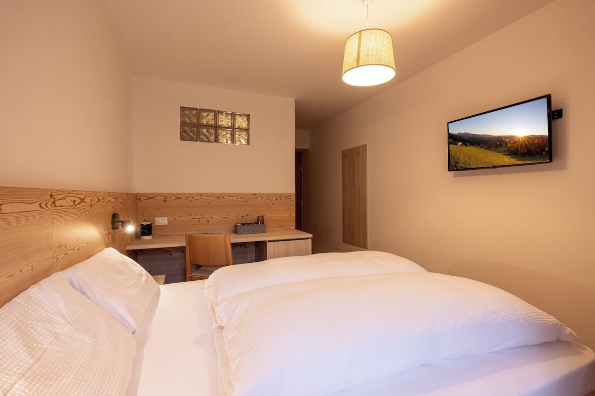 A bedroom with a wooden headboard, a bed with white bedding, a desk with a chair, a lamp, and a television on the wall.