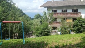 Una casa a due piani con un balcone, circondata da vegetazione lussureggiante e un'altalena in primo piano.