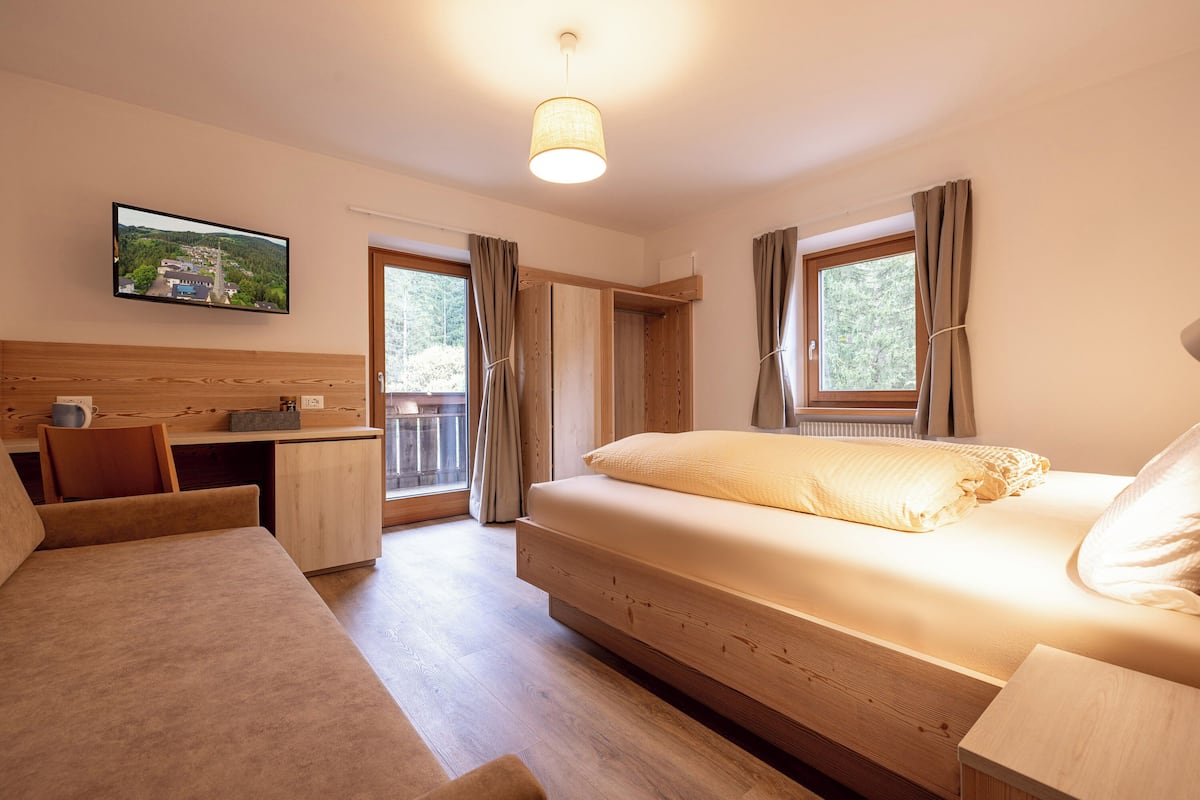 A bedroom with a bed, a desk, a chair, a TV, and a window with curtains.