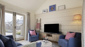 A living room with two blue armchairs, a round coffee table, a flat-screen TV, and a fireplace. - The Stables at Sturt Farm Courtyard - Beautiful dog friendly home in the Cotswolds with a hot tub (Burford)