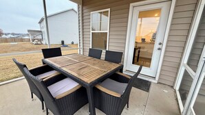 En uteplats med ett träbord och flätade stolar, en skjutdörr i glas och utsikt över ett bostadsområde.