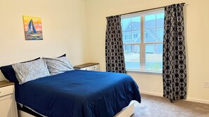 Ein Schlafzimmer mit einem Bett, einem Nachttisch, einem Fenster mit gemusterten Vorhängen und einem Bild an der Wand.