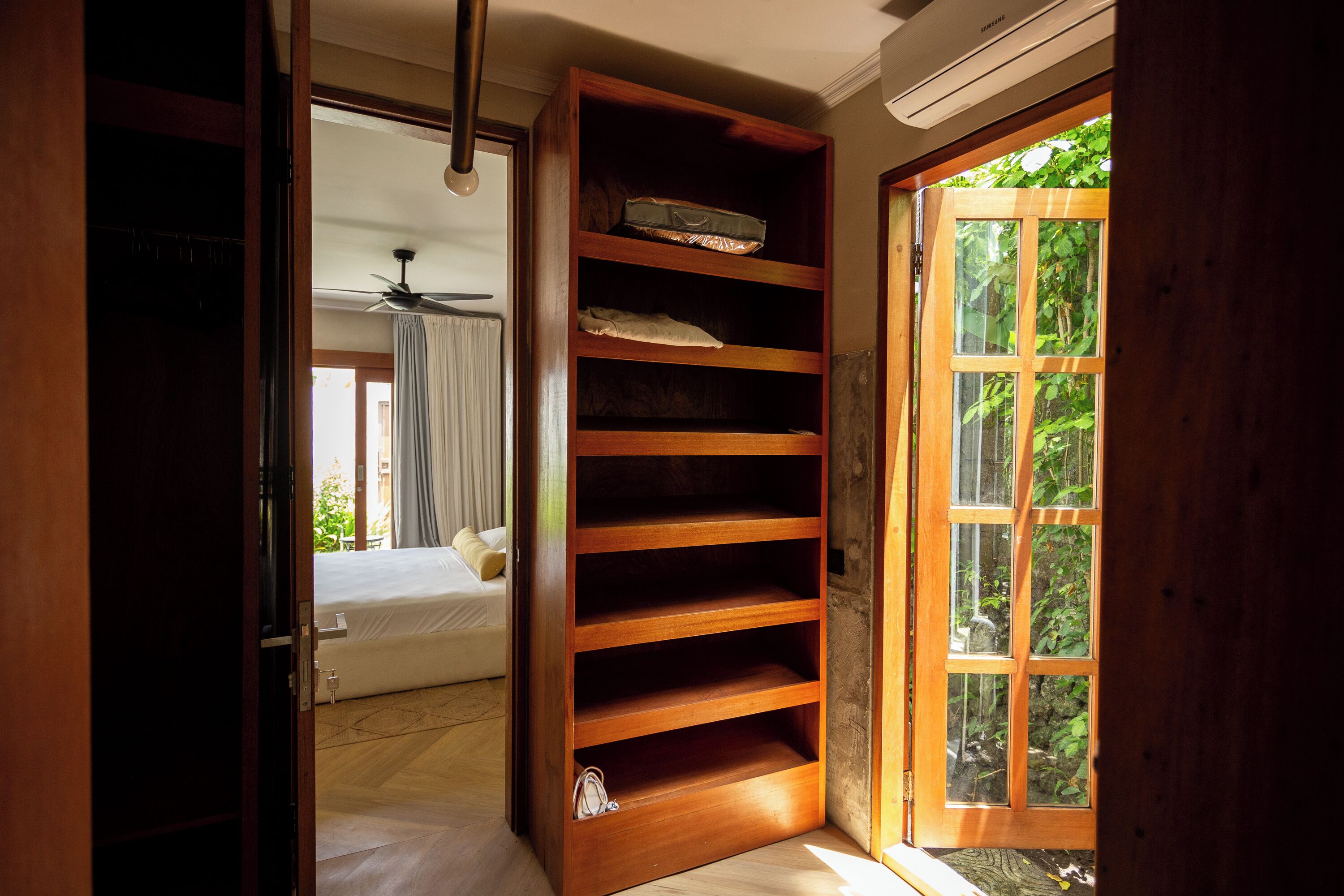 A room with a wooden shelving unit, a bed with a white headboard, and a view of greenery through a glass door.