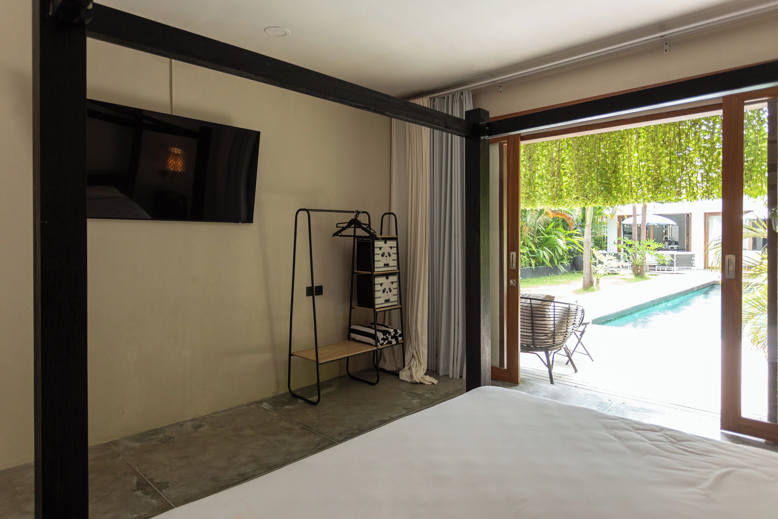 Une chambre dotée d’un grand lit, d’un téléviseur fixé au mur, d’une étagère en bois avec des boîtes de rangement, et offrant une vue sur une piscine et un espace extérieur.