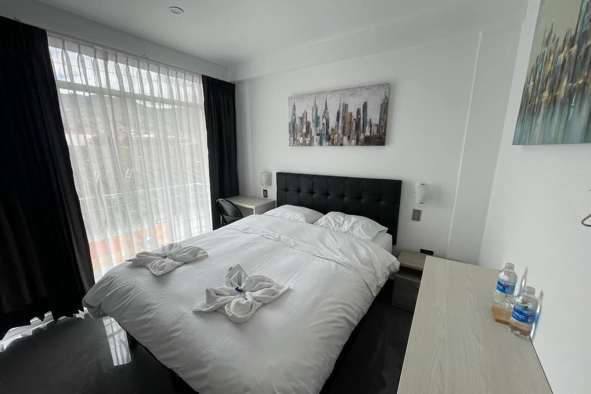 A hotel room with a large bed, a bedside table, a nightstand, and a cityscape painting on the wall.