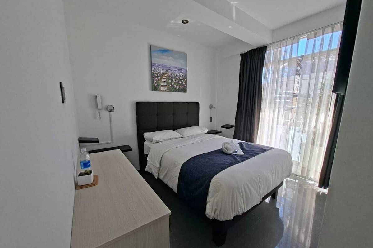 A modern hotel room with a large bed, a desk, a chair, a nightstand, a lamp, and a window with curtains.