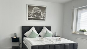 A modern bedroom with a large bed, a nightstand, a framed picture on the wall, and a window with blinds. - Penthouse, KingSize Bed, Queen Size SofaBed, full Kitchen, 650 MBit, Workspace (Euskirchen)