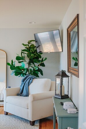 A room with a white armchair, a green side table with a lantern, a potted plant, and a wall-mounted TV. - The Guest Wing at Kateri: Luxury Stay Waynesboro (Waynesboro)