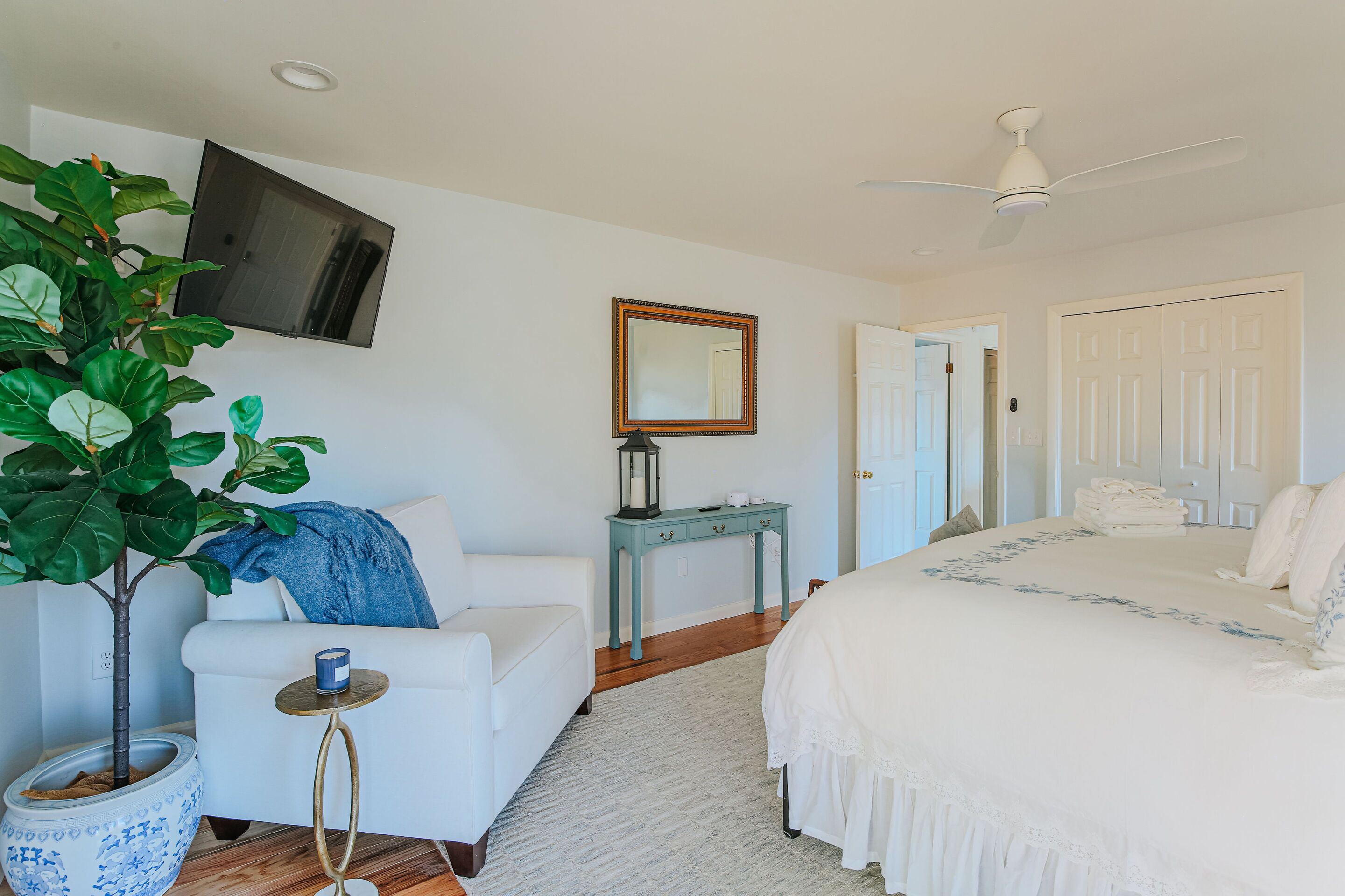 A bedroom with a bed, a chair, a small table, a plant, a mirror, and a ceiling fan.