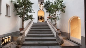 Eine Treppe mit aufwendiger Balustrade, gesäumt von Kübelpflanzen, die zu einem hell erleuchteten Innenraum führt.