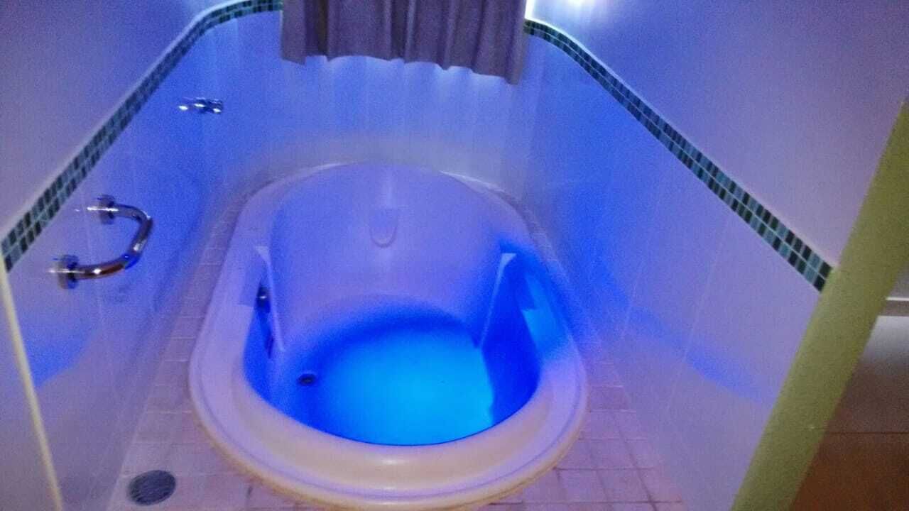 A small, blue-lit toilet stall with a white seat and a metal grab bar.