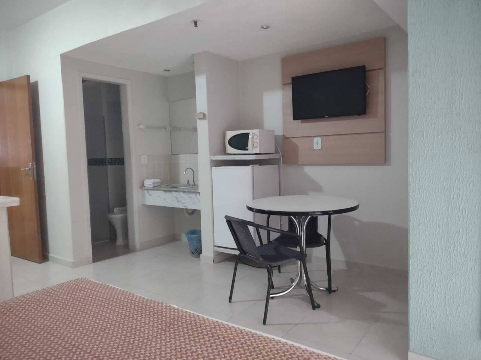 A small, well-lit room with a kitchenette, a TV mounted on the wall, a round table with two chairs, and a door leading to another room.