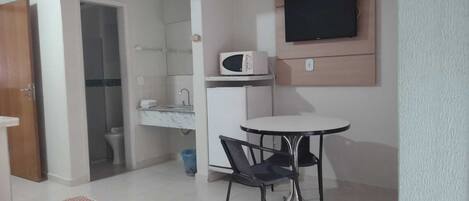 A small, well-lit room with a kitchenette, a TV mounted on the wall, a round table with two chairs, and a door leading to another room.
