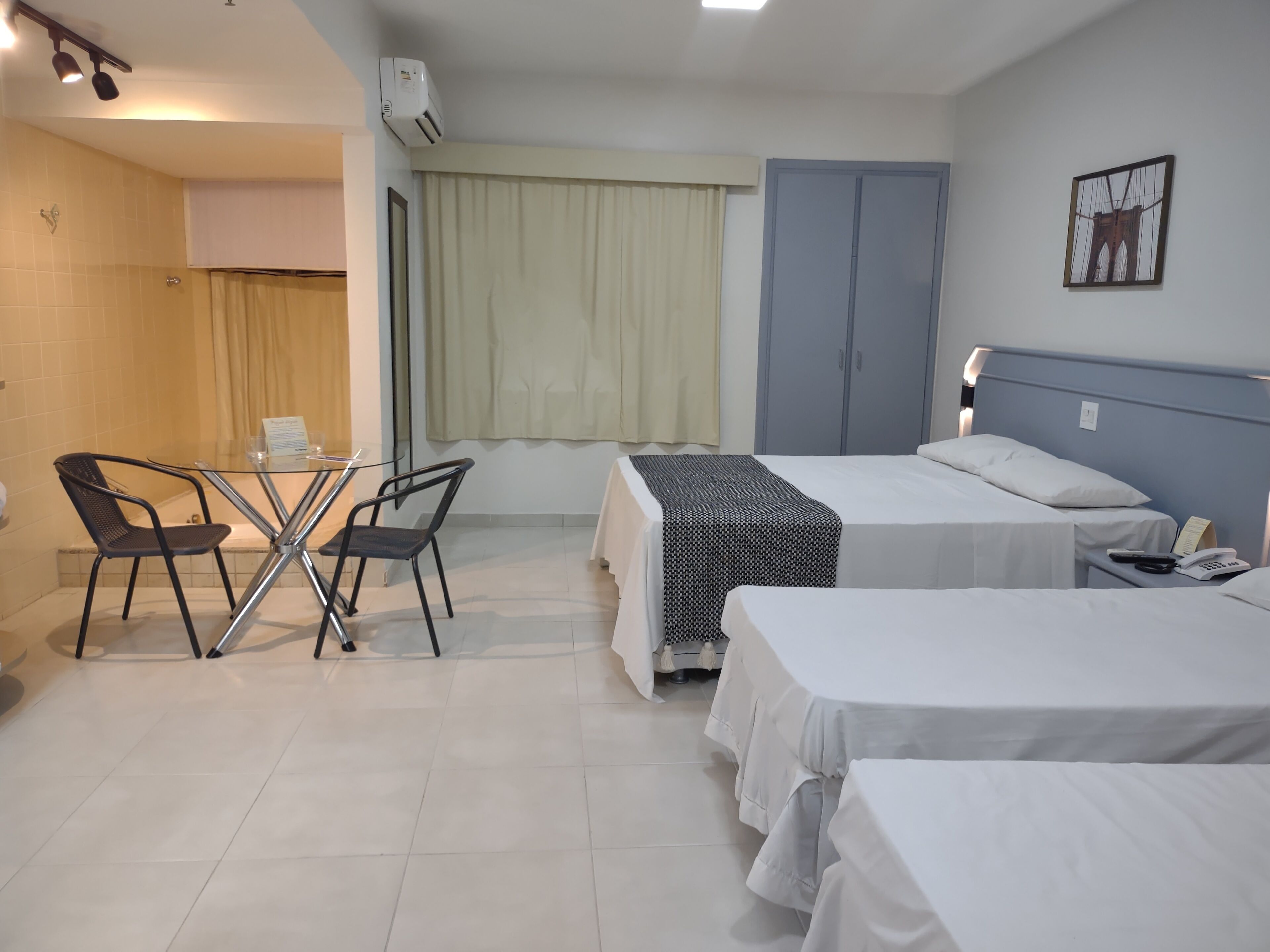 Habitación de hotel con dos camas, una mesita con sillas, una ventana con cortinas, un armario y un cuadro colgado en la pared.