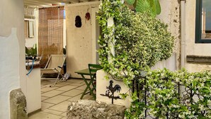 Ein überdachter Patio-Bereich mit einer Steinsitzbank, einem Tisch und einem Stuhl. Eine an der Wand befestigte Hahnschmuck-Figur und ein Topfpflanzen mit weißen Blüten sind ebenfalls vorhanden.