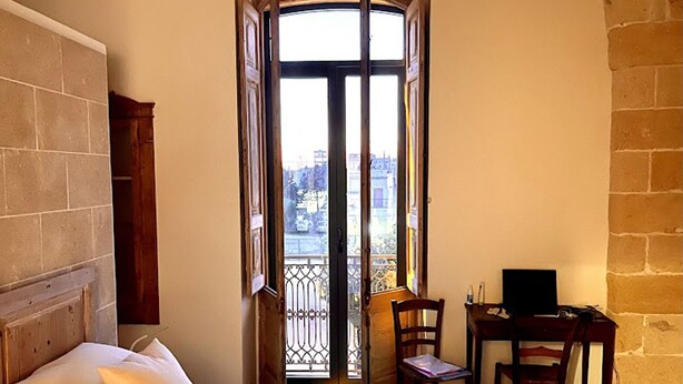 Ein Schlafzimmer mit einem großen Bett, einem Schreibtisch und einem Stuhl, ein Balkon mit Blick auf Gebäude und eine Steinsäule.