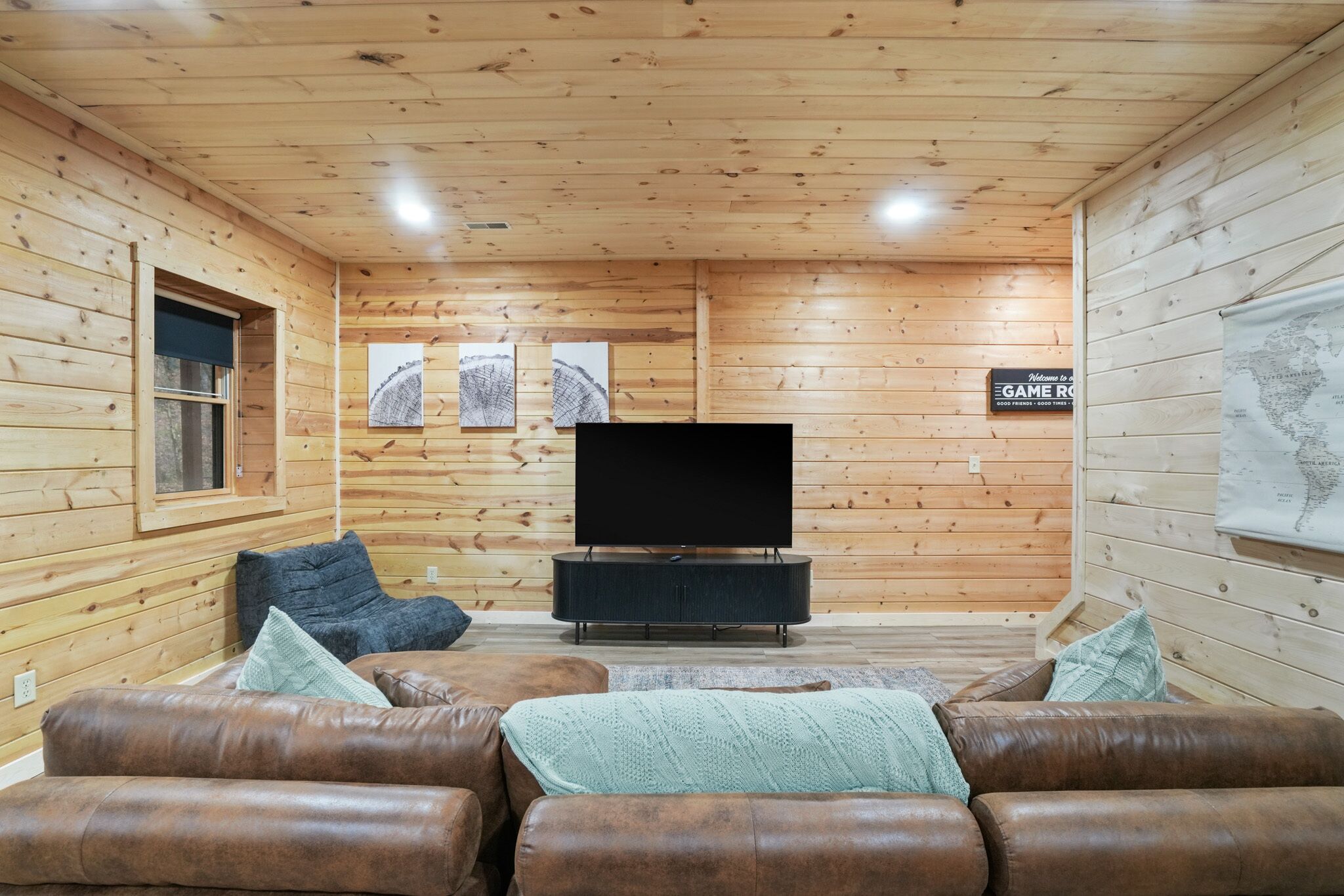 A cozy log cabin interior with a leather sofa set, a flat-screen TV on a black stand, wooden walls, and a map on the right.