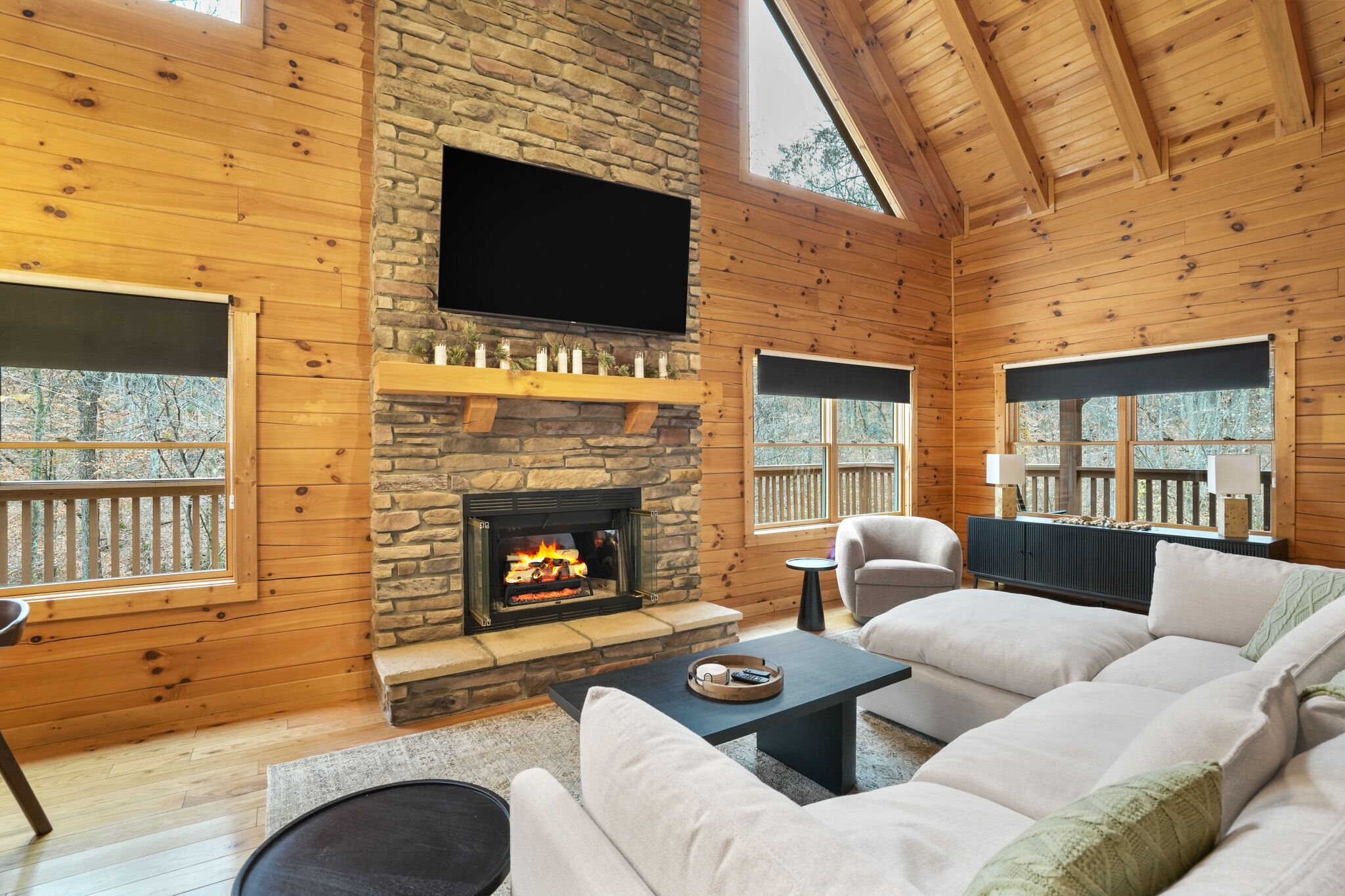 A cozy living room with a stone fireplace, wooden walls, and a large TV mounted above it. There are comfortable sofas and a small table with a tray.