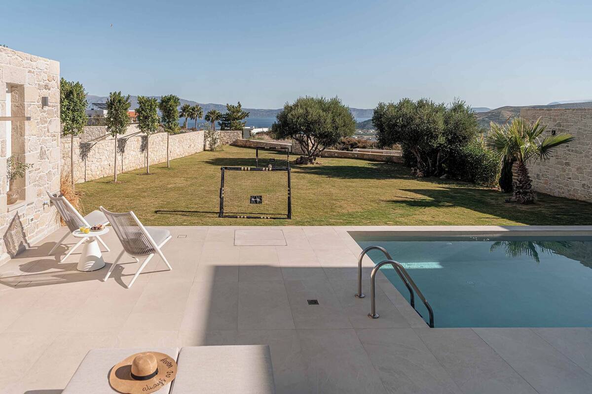 A poolside area with a view of the sea, a stone wall, and a grassy lawn with trees.