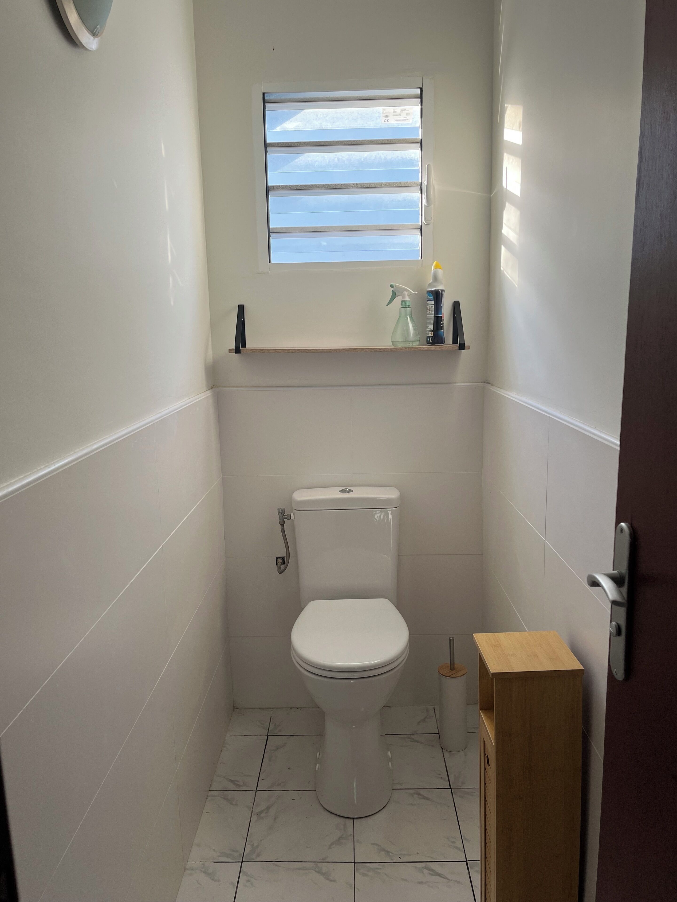 Un bagno piccolo e pulito con il water, una finestra e uno scaffale con prodotti per la pulizia.