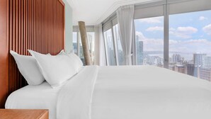 Une chambre d’hôtel avec un grand lit, une tête de lit en bois et une vue sur la ville grâce à de grandes fenêtres.