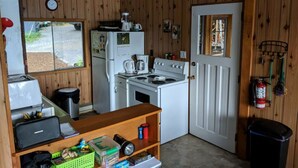 A compact kitchen with wooden walls, a white refrigerator, stove, and sink. There's a window with a view of a driveway, a door with a small window, and a shelf with various items. - Oars n Smores
A place to call yours. (Georgian Bay)