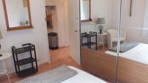 A hotel room with a bed, a small bedside table with a lamp, a chair, a mirror, and a trash bin.