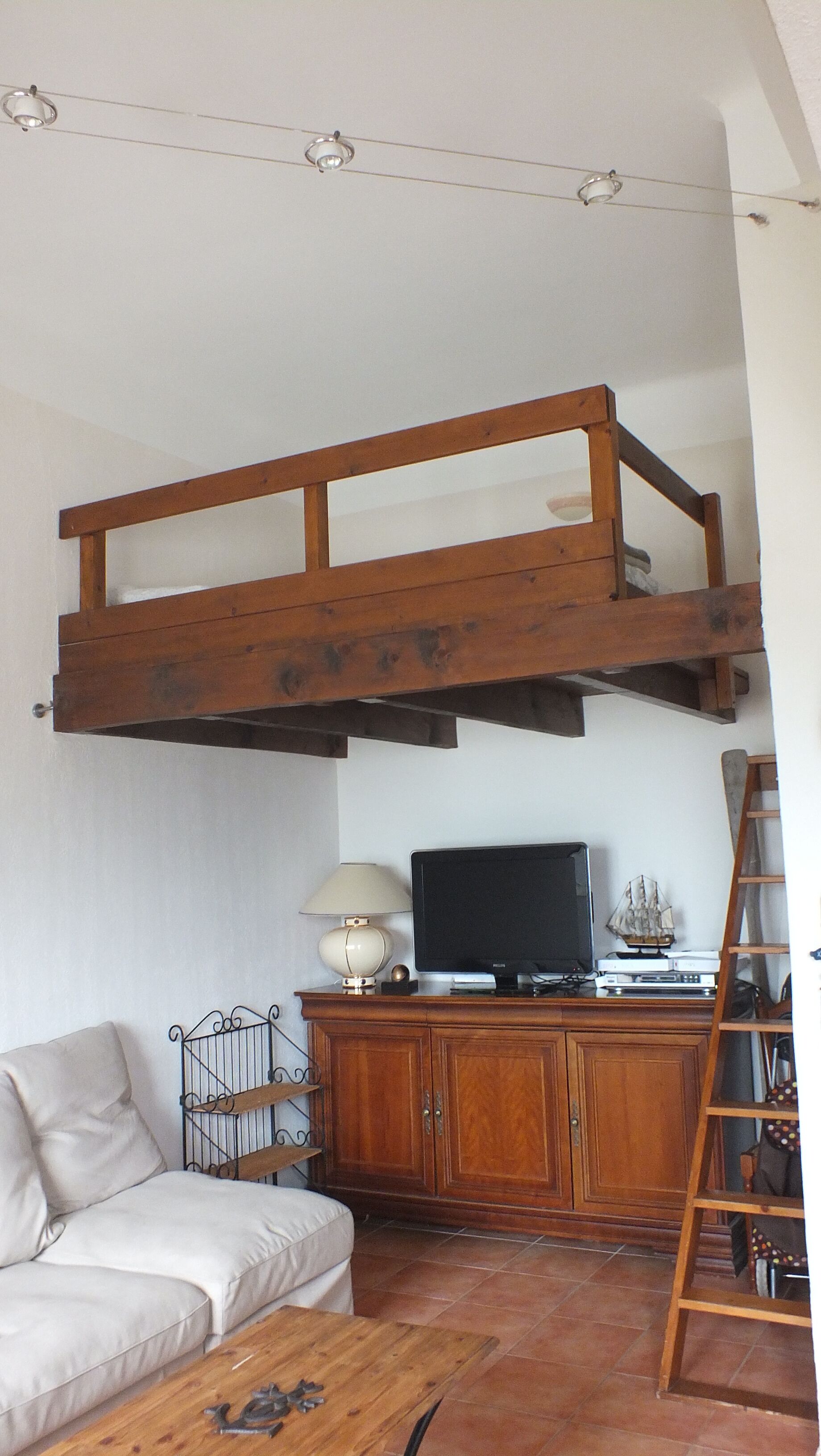Une chambre à coucher aménagée dans un espace ouvert, avec un lit superposé en bois, un téléviseur posé sur une commode en bois, un canapé blanc et une table basse en bois.
