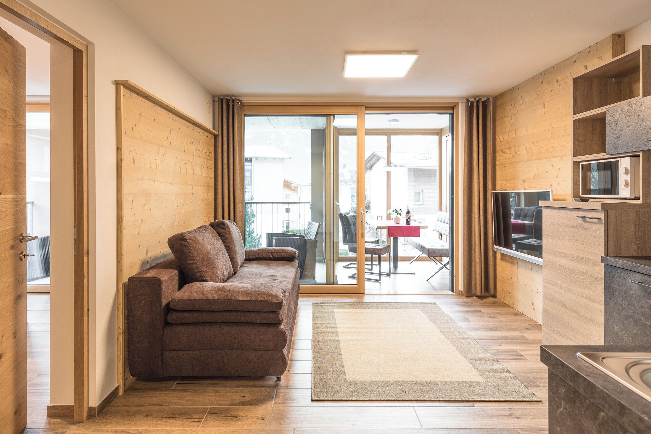 A modern living room with a sofa, a flat-screen TV, wooden flooring, and a sliding glass door leading to an outdoor area.