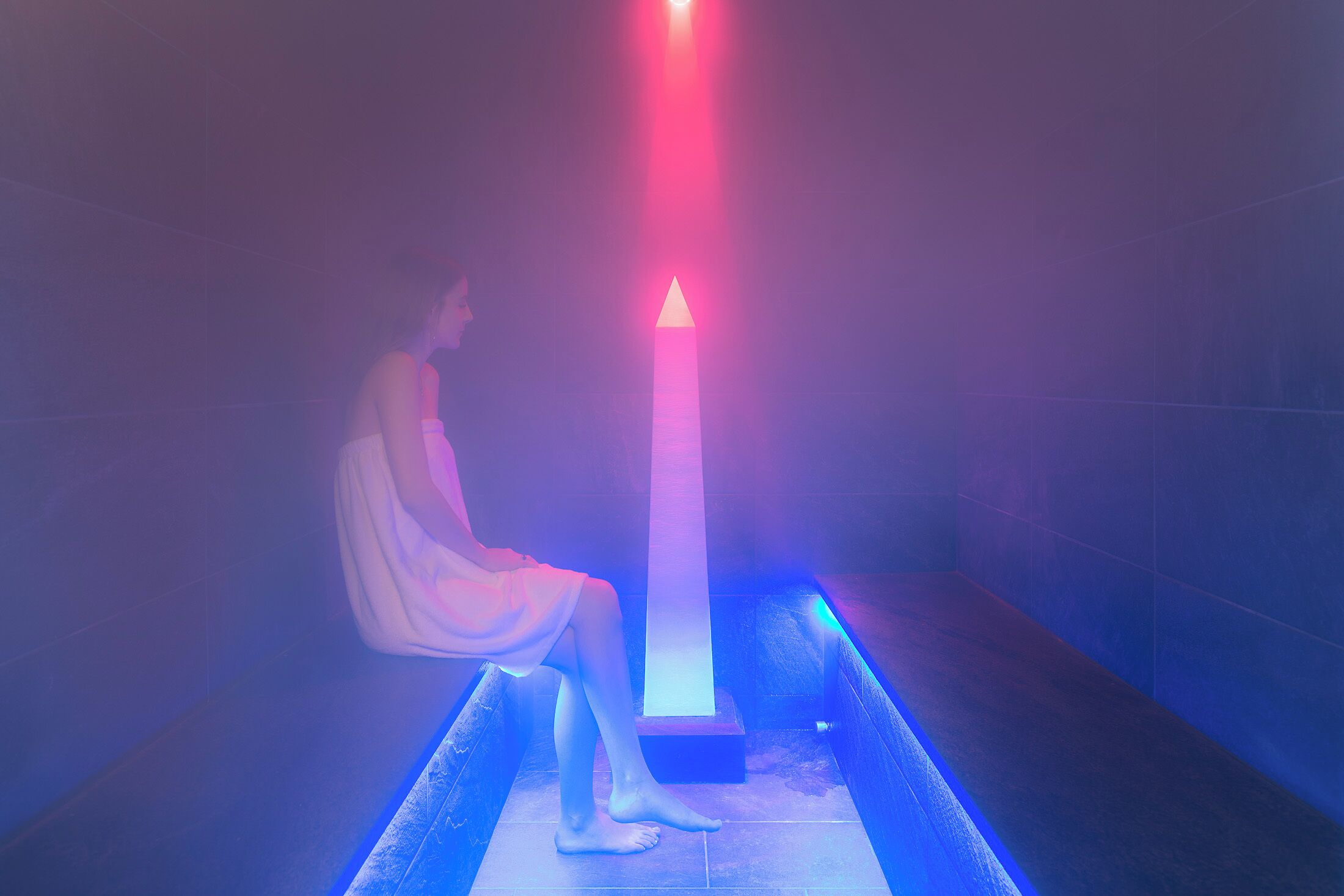 Une personne assise sur un banc dans une pièce faiblement éclairée, avec une source de lumière verticale.