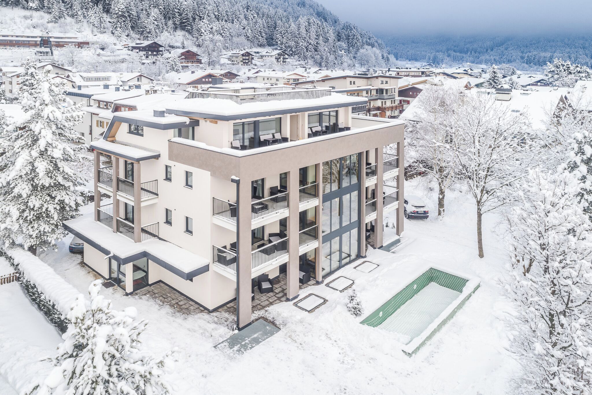 A modern multi-story building with balconies and large windows, surrounded by snow-covered trees and a mountainous backdrop.