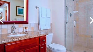 Een badkamer met een wastafel met een granieten blad, een rode wastafelmeubel, een wit toilet, een douchecabine van glas en een grote spiegel.