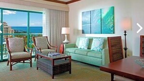 Hotelkamer met groot raam dat uitzicht geeft op de oceaan, een plafondventilator, een groene bank en een houten bijzettafeltje.