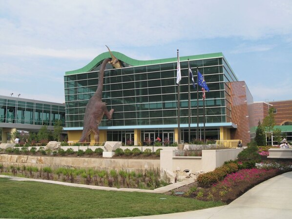 A modern building with a glass facade, a large dinosaur sculpture in front, and flags including the EU flag. - Private Pickleball Court+Huge Fenced Yard, Easy Access to Grand Park & Downtown (Indianapolis)