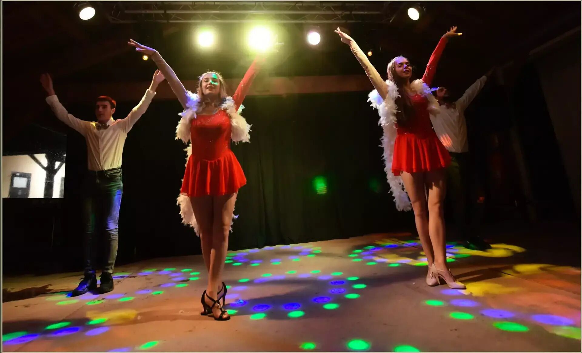 Un palco illuminato da luci da palcoscenico, con tre ballerine in abiti rossi e costumi piumati che si esibiscono.