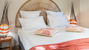 Un letto con testiera intrecciata, cuscini bianchi e un cuscino decorato con motivi floreali rosa. Su entrambi i lati del letto, due lampade alte e slanciate con strisce arancioni e bianche.