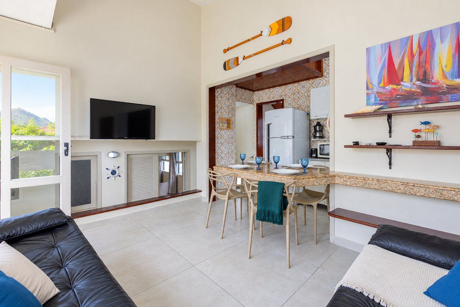 Uma sala moderna com televisão de tela plana, uma área de jantar com cadeiras e mesa, e uma cozinha com geladeira e micro-ondas.